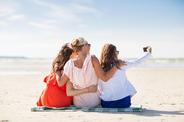 vacances d'été, vacances, voyage, technologie et concept de personnes - groupe de jeunes femmes souriantes prenant selfie avec smartphone sur la plage