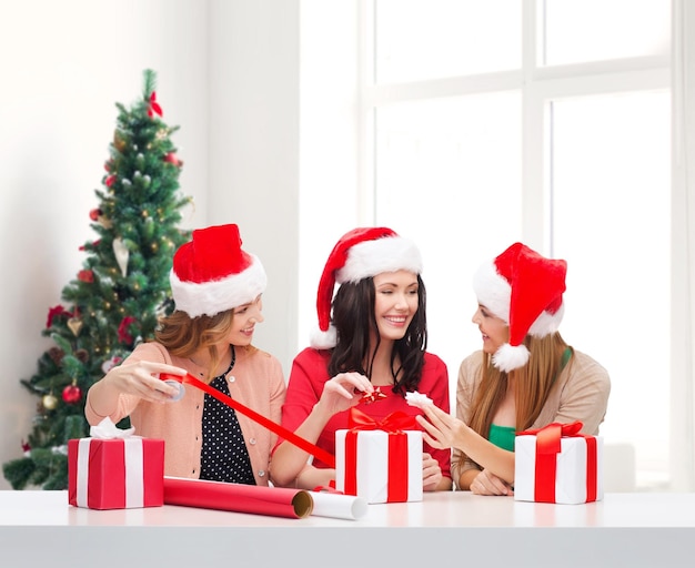 vacances, fête, décoration et concept de personnes - femmes souriantes en chapeaux de santa helper avec papier de décoration et coffrets cadeaux sur fond de salon et d'arbre de noël