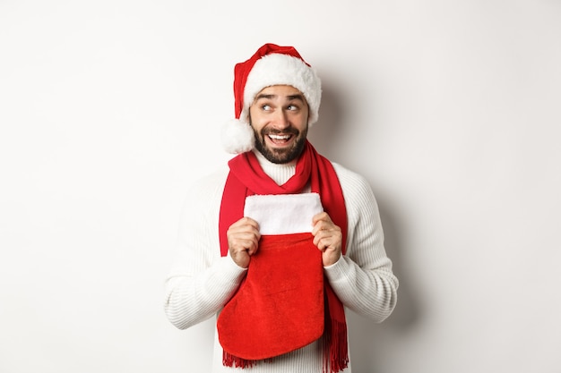 Vacances d'hiver et concept de shopping. Un bel homme excité planifiant le nouvel an, tenant une chaussette de Noël pour des cadeaux et pensant, debout sur fond blanc