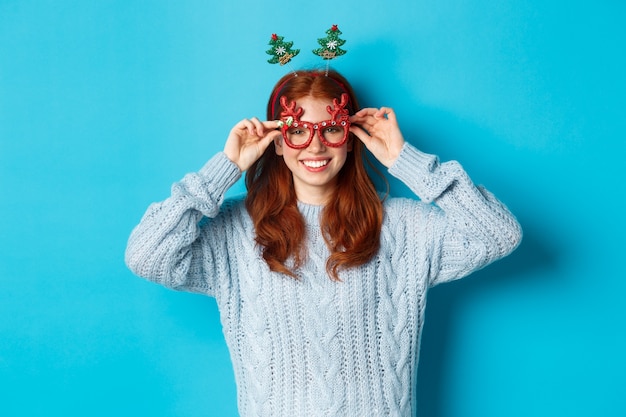 Vacances d'hiver et concept de vente de Noël. Beau modèle féminin rousse célébrant le Nouvel An, portant un bandeau et des lunettes de fête drôle, souriant à la caméra.