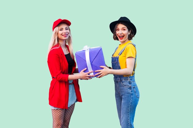 Vacances lumineuses de charmants amis. Portrait de deux beaux hipsters à la mode heureux, fille donnant à son meilleur ami une grande boîte violette présente, regardant la caméra. Studio shot, isolé sur fond vert