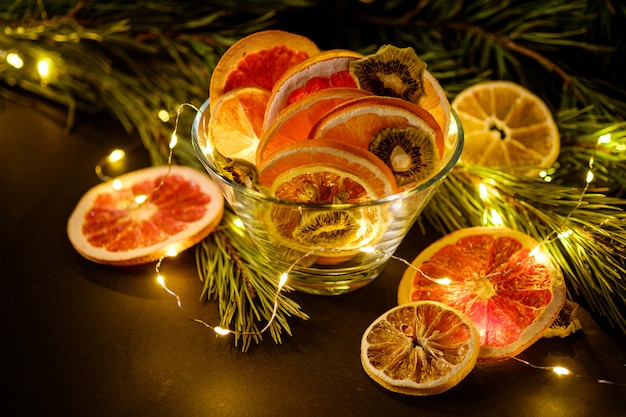 Vacances de mauvaise humeur créative Noël fruits du Nouvel An avec pamplemousse séché, kiwi, orange et citron dans un bol en verre avec une branche de sapin avec des lumières LED chaudes, vue d'angle