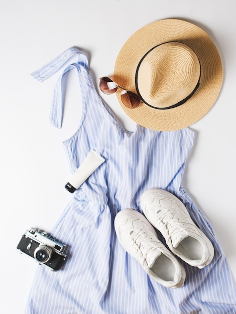 Vacances de mode à plat avec robe d'été en paille bleue chapeau baskets lunettes de soleil et autres accessoires sur fond blanc vue de dessus verticale