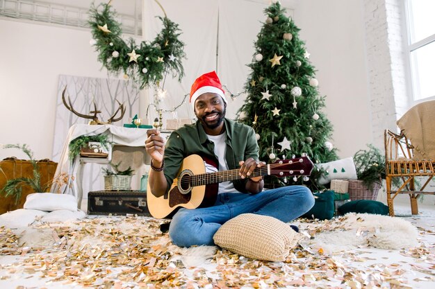 Vacances de Noël Concept du Nouvel An Un homme africain souriant et tenant la lumière du Bengale et la guitare est assis sur le sol dans un salon confortablement décoré