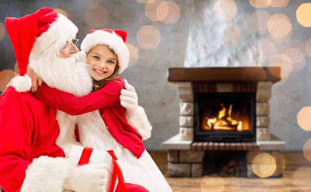 vacances, noël, hiver et concept de personnes - fille heureuse avec le père noël et boîte-cadeau sur fond de cheminée et de lumières