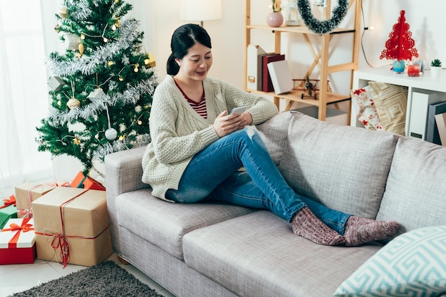 vacances de noël insouciantes femme coréenne du millénaire se relâchant sur le canapé en parcourant les messages sur la plate-forme de médias sociaux. technologie et concept de style de vie authentique.