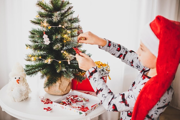 Vacances de Noël et nouvel an. Garçon enfant décorant le petit arbre de Noël pendant les vacances d'hiver. Adolescent en pyjama et bonnet de Noel rouge suspendu à un jouet en bois décoratif sur des branches de sapin.