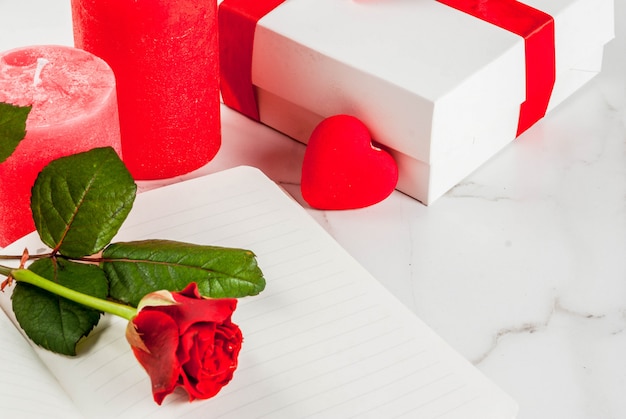 Vacances, Saint Valentin. Bouquet de roses rouges, cravate avec un ruban rouge, avec bloc-notes vierge, coffret cadeau enveloppé et bougie rouge. Sur une table en marbre blanc, vue de dessus du fond