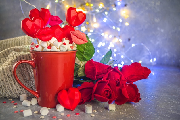 Vacances de la Saint-Valentin avec tasse de chocolat chaud rouge et ensemble de bonbons coeurs rouges sur des bâtons en tasse, avec des roses rouges et effet de lumières bokeh