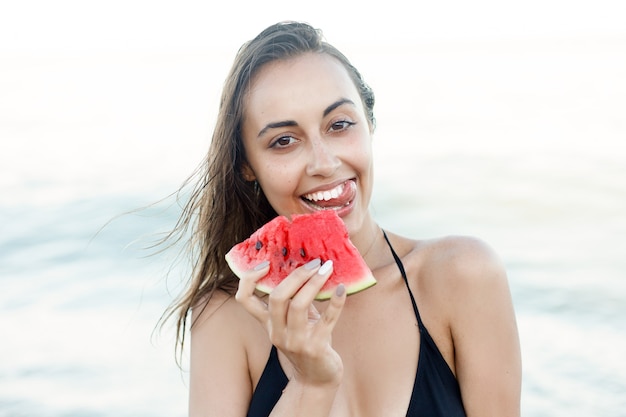 Vacances, station balnéaire, concept touristique - Vacances d'été - jeune fille mangeant de la pastèque fraîche sur la plage de sable. belle jeune femme mange de la pastèque sur la plage aux chaudes journées d'été.