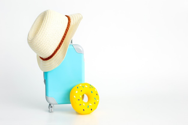 Une valise bleue sur roues un chapeau de paille et un beignet gonflable jaune se tiennent sur un fond blanc