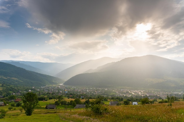 Vallée de la beauté avec village et montagnes autour sous la pluie d'été des nuages sombres au coucher du soleil