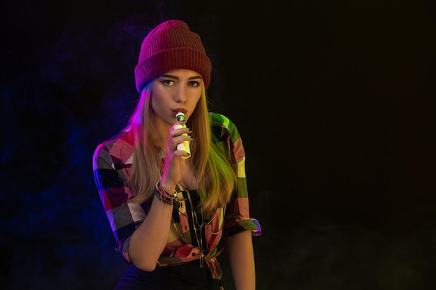 Vapoteuse. Jeune femme hipster vape e-cig sur fond noir. Style hip-hop. Notion de vapeur. Vapoter une e-cigarette. Prise de vue en studio