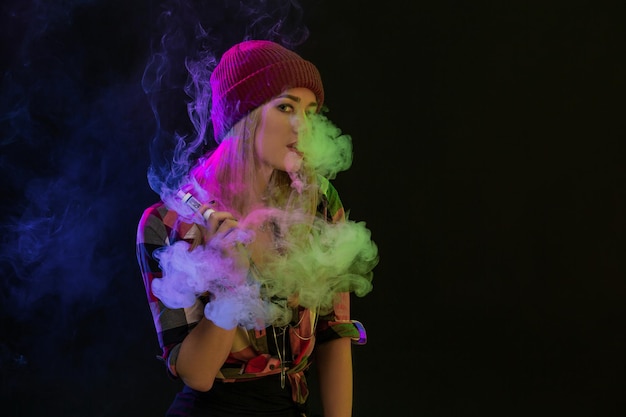 Vapoteuse. Jeune femme hipster vape e-cig sur fond noir. Style hip-hop. Notion de vapeur. Vapoter une e-cigarette. Prise de vue en studio