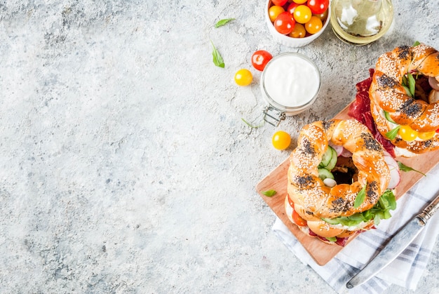 Variété de sandwichs bagels faits maison avec sésame et graines de pavot, fromage à la crème, jambon, radis, roquette, tomates cerises, concombres, surface texturée gris blanc vue de dessus