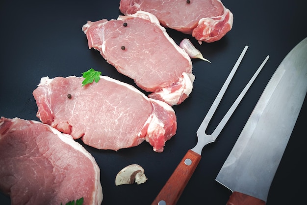 Variété de steaks de viande de boeuf cru avec des ingrédients pour cuisiner un couteau et une fourchette à viande sur fond sombre