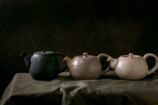 Variété de théières artisanales en céramique faites à la main bouilloires en rangée pour la cérémonie du thé debout sur une nappe en lin dans une pièce sombre.