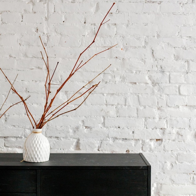 Un vase en céramique blanche avec des branches sèches sur un socle noir contre un mur de briques blanches Intérieur
