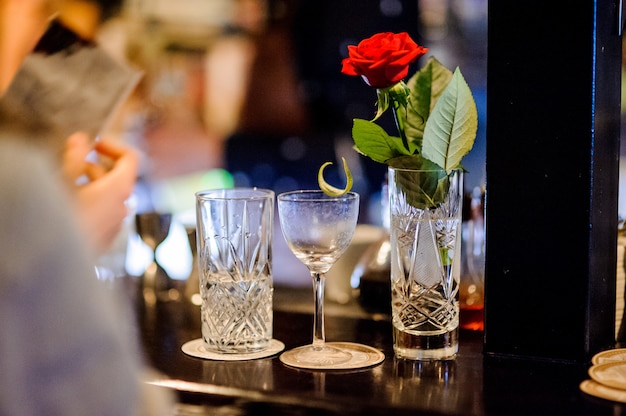 Vase en cristal avec rose rouge, un verre vide et un verre à moitié rempli d'eau sur un comptoir de bar