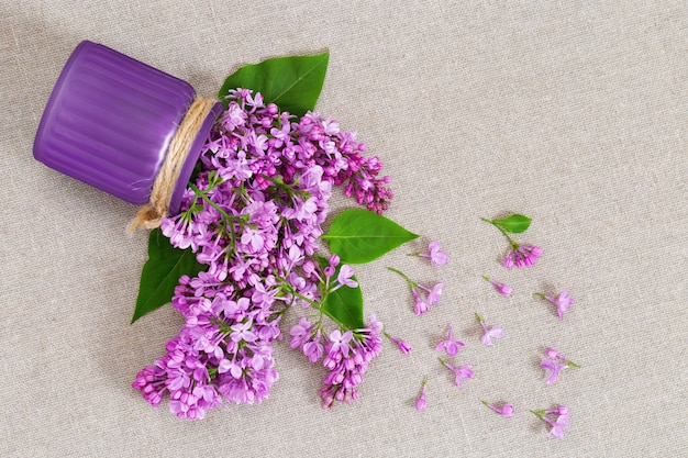 Vase avec des fleurs lilas éparpillées sur un matériau brut naturel.