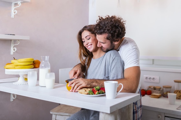 Les végétariens féminins et masculins se tiennent ensemble contre l'intérieur de la cuisine, préparent une salade de fruits. Un couple de famille cuisine dans une cuisine confortable, le mari aide la jeune femme à préparer le petit-déjeuner pour toute la famille.