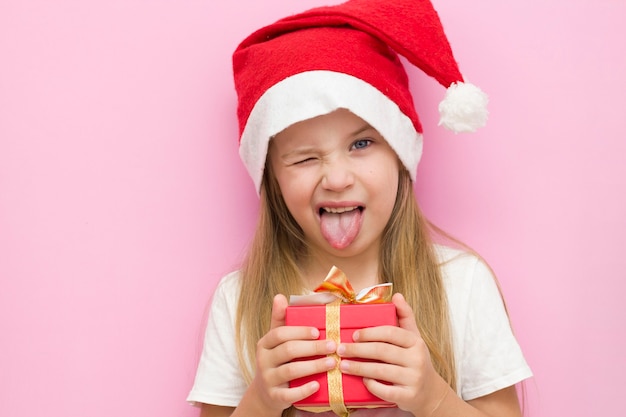 La veille de Noël! Un enfant joyeux dans un chapeau de Noël sur fond rose avec une boîte cadeau rouge.