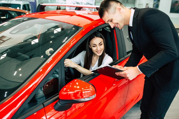 Le vendeur du concessionnaire montre une tablette avec des documents pour se familiariser avec une belle fille souriante assise à l'intérieur d'une voiture rouge