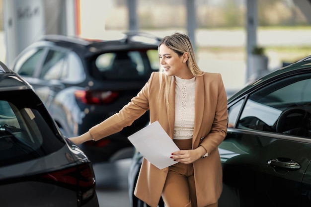 Un vendeur heureux du centre de concession automobile tient un contrat entre ses mains et touche la voiture dans la salle d'exposition