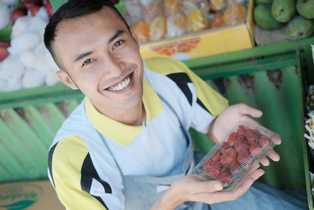 Vendeur masculin souriant portant un paquet de fraises à deux mains dans un magasin de fruits