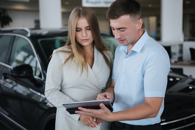 Vendeur parlant avec une femme et aidant à choisir une nouvelle voiture homme et femme dans une salle d'exposition de voitures homme utilisant