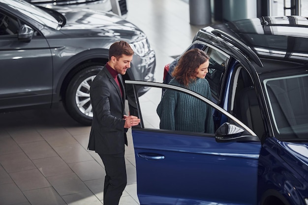 Vendeur professionnel aidant une jeune fille en choisissant une nouvelle automobile moderne à l'intérieur.