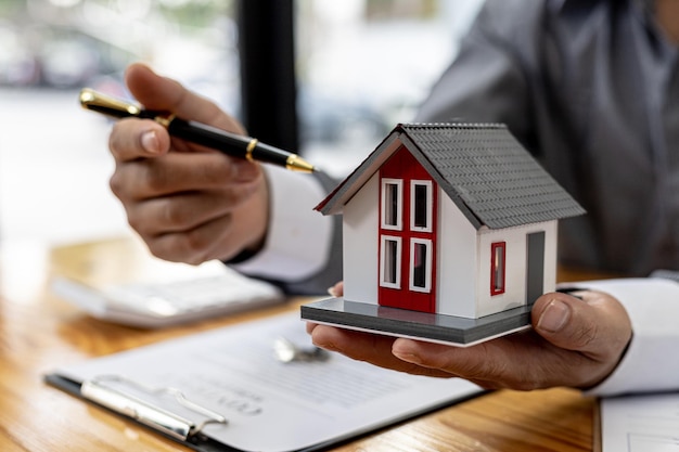 Vendeur tenant une maquette orange d'une petite maison, un vendeur d'un projet de logement rédige un contrat de vente pour un client qui réserve une maison dans un projet qu'il entretient. Concept de commerce immobilier.