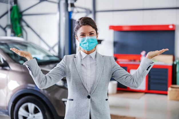 Vendeur de voiture femme souriant et sympathique avec masque facial debout dans le garage du salon de voiture et montrant autour du garage. La voiture est prête et réparée.