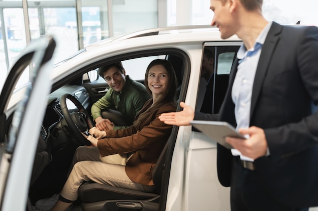 Vendeur de voitures du millénaire discutant de l'achat d'une nouvelle voiture avec un jeune couple heureux après un essai routier à