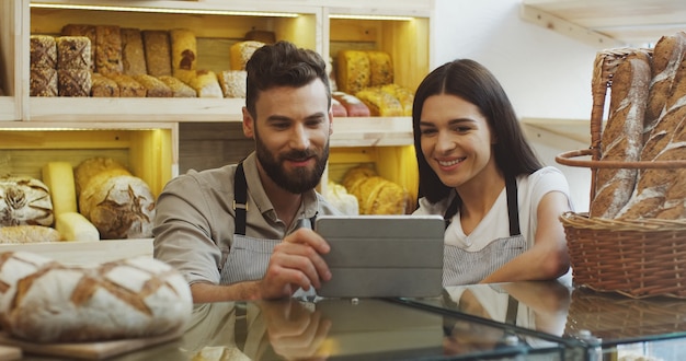 Vendeurs de pain masculins et féminins attrayants faisant défiler et regarder quelque chose sur la tablette tout en se tenant au comptoir de la boulangerie. Intérieur