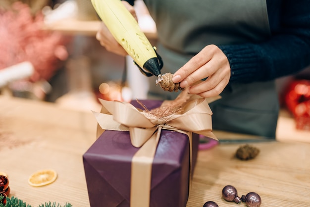 Une vendeuse décore une boîte-cadeau avec un cône de sapin, un processus d'emballage et de décoration à la main. Femme enveloppe présente sur la table, procédure de décoration