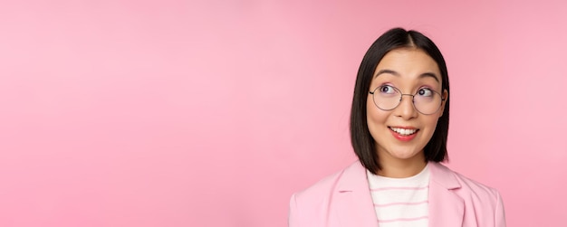 Vendeuse enthousiaste femme d'affaires asiatique dans des verres regardant intriguée par la publicité de l'entreprise du logo du coin supérieur gauche debout sur fond rose