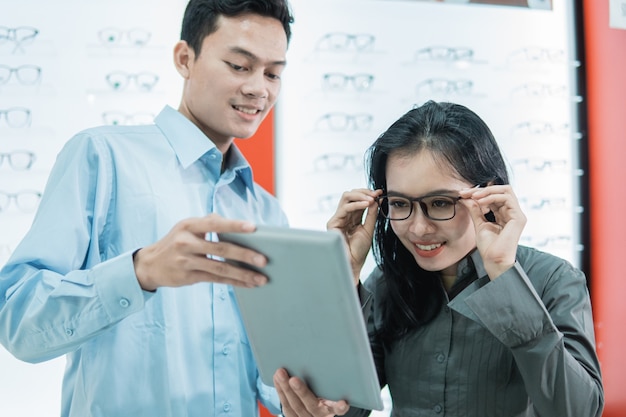 Une vendeuse servant des clientes essayant des lunettes pour regarder l'écran de la tablette en se tenant debout dans le contexte d'une vitrine de support de lunettes chez un opticien