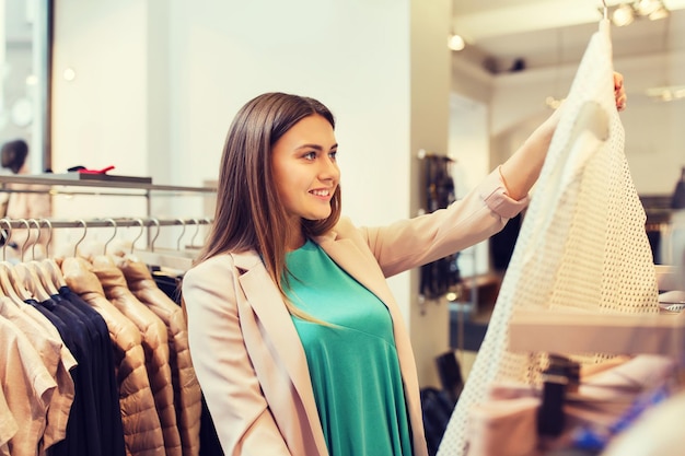 vente, shopping, mode, style et concept de personnes - jeune femme heureuse choisissant des vêtements dans un centre commercial ou un magasin de vêtements