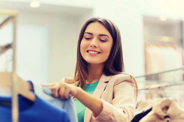 vente, shopping, mode, style et concept de personnes - jeune femme heureuse choisissant des vêtements dans un centre commercial ou un magasin de vêtements