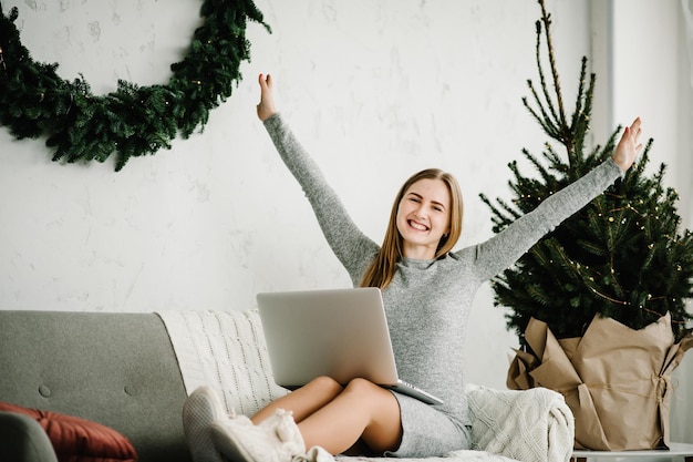 Ventes de Noël Jeune femme utilisant un ordinateur portable et travaillant au bureau à domicile près d'un pin décoré avec un intérieur de Noël Fille tapant sur un ordinateur portable à la maison Concept de Noël Planification des vacances Recherche Web