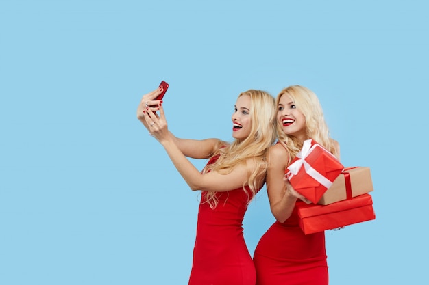 Ventes de vacances. Femmes heureuses avec des coffrets cadeaux en robes rouges, fait selfie sur smartphone.