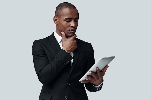 Vérification de chaque détail. Jeune homme africain en tenue de soirée travaillant à l'aide d'une tablette numérique en se tenant debout sur fond gris