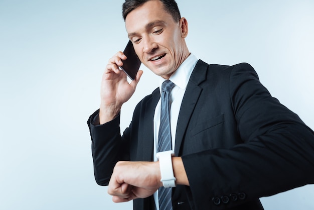 Vérification du temps. Agréable bel homme d'affaires occupé en regardant sa smartwatch et en vérifiant l'heure tout en parlant au téléphone