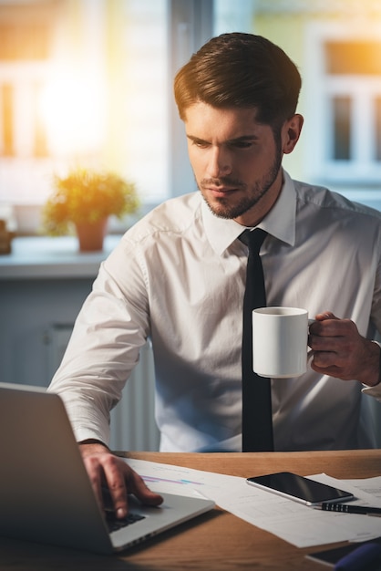 Vérification de son courrier électronique. Jeune bel homme pensif utilisant son ordinateur portable et tenant une tasse de café