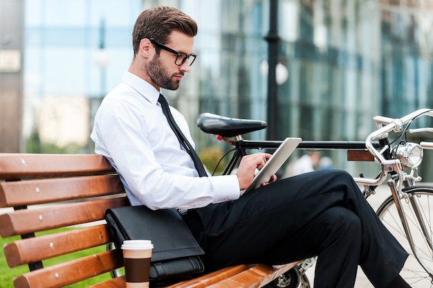 Vérification de son emploi du temps. Vue latérale d'un jeune homme d'affaires confiant travaillant sur une tablette numérique tout en étant assis sur le banc près de son vélo avec un immeuble de bureaux en arrière-plan