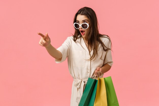 Vérifiez cela, doit avoir cette saison. Étonné et excité jeune shoppaholic femme enthousiaste, voyant une chose incroyable dans l'allée, tenant des sacs d'expédition et pointant le doigt vers la gauche, mur rose