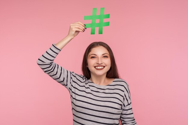 Vérifiez le sujet Internet viral. Portrait d'une jeune femme heureuse en sweat-shirt symbole hashtag au-dessus de la tête et regardant la caméra avec un sourire à pleines dents, suivant les tendances. tourné en studio isolé sur fond rose