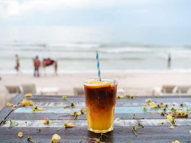 Un verre d'americano orange glacé frais décoré de tranches d'orange servi sur une table en bois sur la plage vue sur le paysage marin Café noir froid avec du jus d'orange mélangeant un menu de boissons sans alcool