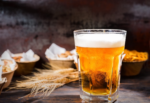 Verre avec de la bière fraîchement versée près des assiettes avec pistaches, petits bretzels et cacahuètes sur un bureau en bois foncé. Concept de nourriture et de boissons